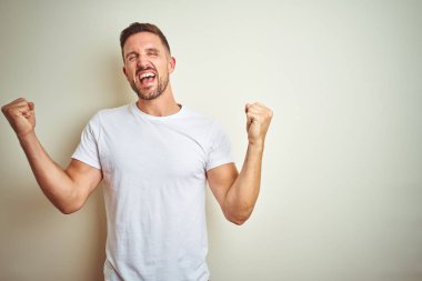 İzole edilmiş arka plan üzerine gündelik beyaz tişört giyen genç yakışıklı adam kolları havada ve gözleri kapalı olarak başarının şaşkınlığını ve şaşkınlığını kutluyor. Kazanan konsept.