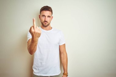 İzole arka plan gösteren orta parmak, kaba ve kaba siktir git ifade üzerinde rahat beyaz t-shirt giyen genç yakışıklı adam