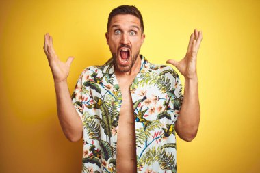 Sarı izole arka plan üzerinde yaz hawaii çiçek gömlek giyen yakışıklı adam deli kutluyor ve kollarını kaldırdı ve açık gözleri heyecan çığlık ile başarı için hayran. Kazanan kavramı