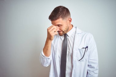 İzole arka planda beyaz kürk giyen yakışıklı doktor yorgun burnu ve gözleri ovuşturup yorgun ve baş ağrısı çekiyor. Stres ve hüsran kavramı.