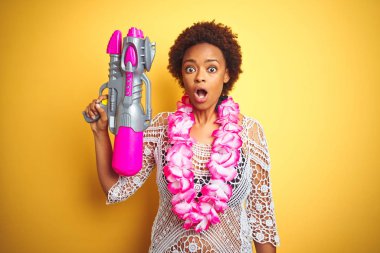Afro saç giyen çiçek Hawaii Lei ve su tabancası ile genç Afrikalı Amerikalı kadın sürpriz bir yüz ile şok korkuyor ve korku ifadesi ile heyecanlı korkmuş