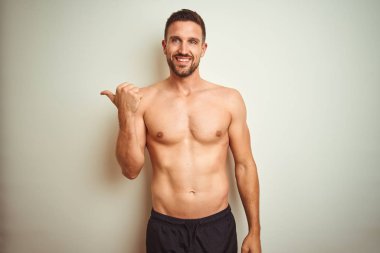 Genç, yakışıklı, üstsüz, soyutlanmış arka planda gülümseyen, gülümseyen ve baş parmağıyla yan tarafı gösteren bir adam..