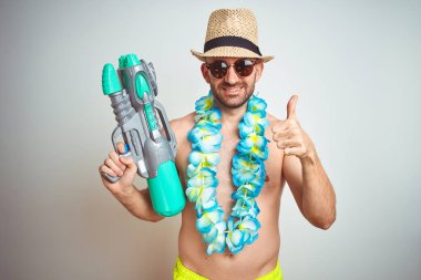 Yaz şapkası takan ve elinde su tabancası olan genç bir adam izole edilmiş arka planda mutlu bir şekilde gülümsüyor.