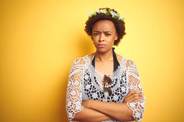 Young african american woman with afro hair wearing flowers crown over yellow isolated background skeptic and nervous, disapproving expression on face with crossed arms. Negative person.