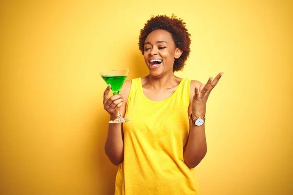 Young african american woman with afro hair drinking a cocktail over yellow isolated background very happy and excited, winner expression celebrating victory screaming with big smile and raised hands