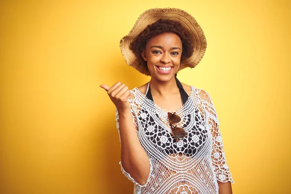 Young african american woman with afro hair wearing summer hat over white isolated background smiling with happy face looking and pointing to the side with thumb up.