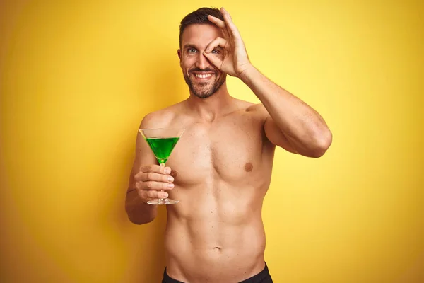 Young handsome shirtless man drinking a summer cocktail over isolated yellow background with happy face smiling doing ok sign with hand on eye looking through fingers