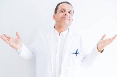 Orta yaş doktor adam beyaz arka plan üzerinde tıbbi ceket giyen habersiz ve kol lar ve eller kaldırdı ile karışık ifade. Şüphe kavramı.