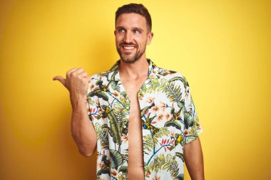 Sarı izole arka plan üzerinde yaz hawaii çiçek gömlek giyen Yakışıklı adam mutlu yüz görünümlü gülümseyerek ve başparmak ile yan işaret.