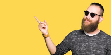 Yüzünde, kocaman bir gülümseme ile güneş gözlüğü takmış genç hipster adam kameraya bakarak yan el ve parmak ile işaret.