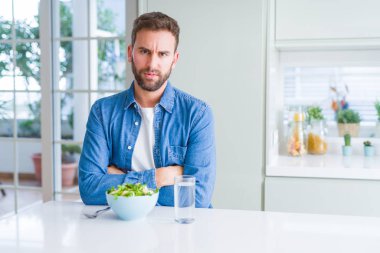 Yakışıklı adam taze sağlıklı salata yeme şüpheci ve sinir, çapraz kolları ile yüzünde onaylamayan ifade. Negatif kişi.