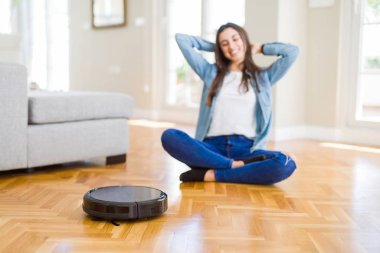 Yerleri temizlemek için otomatik süpürge kullanan genç bir kadın makine ev işi robotu kontrol ediyor.