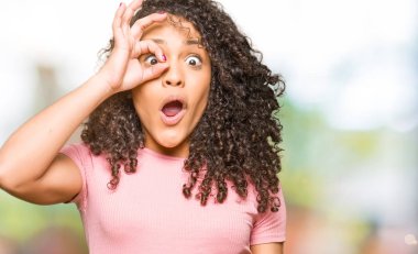 Pembe tişört giyen, kıvırcık saçlı genç bir kadın. Şok olmuş, şaşırmış bir yüz ve parmaklarından bakan bir göz hareketi yapıyor. İnançsız ifade.