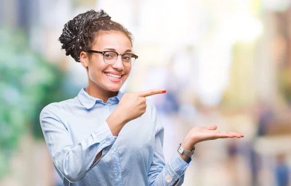Young braided hair african american business girl wearing glasses over isolated background amazed and smiling to the camera while presenting with hand and pointing with finger.