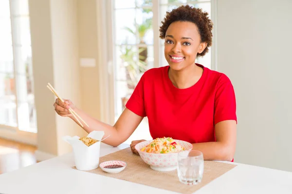Young african american woman with afro hair eating asian food at home with a happy face standing and smiling with a confident smile showing teeth