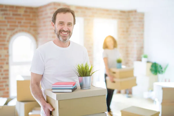 Middle age senior couple moving to a new house, man smiling happy in love with new apartment and holding cardboard boxes