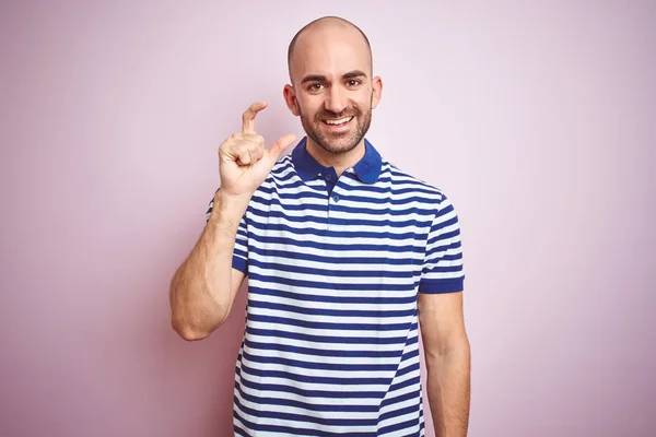 ピンクの孤立した背景の上にカジュアルなストライプの青いTシャツを着てひげを生やした若いハゲ男は 指を見て カメラで小さなサイズのサインをやって手で自信を持ってジェスチャー 測定コンセプト — ストック写真