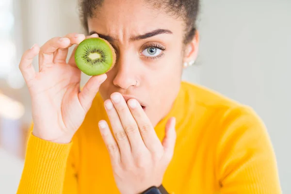 Young african american girl eating green kiwi cover mouth with hand shocked with shame for mistake, expression of fear, scared in silence, secret concept