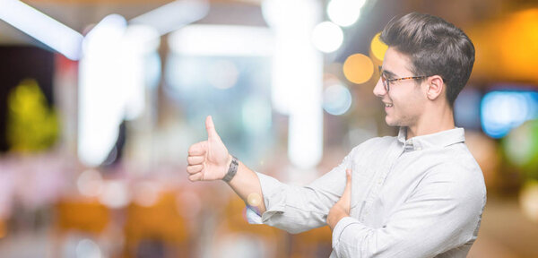 Young handsome man wearing glasses over isolated background Looking proud, smiling doing thumbs up gesture to the side