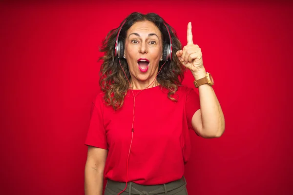 中年のシニア女性は 成功したアイデアで指を指し 赤い孤立した背景の上に音楽を聴いてヘッドフォンを着用しています 終了し ナンバーワン — ストック写真