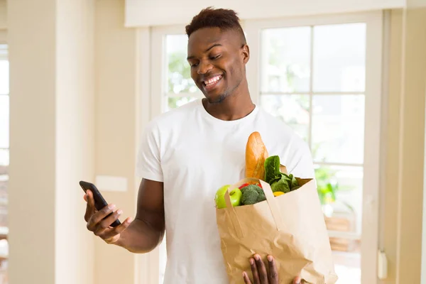 African man holding a paper bag full of groceries and using smar