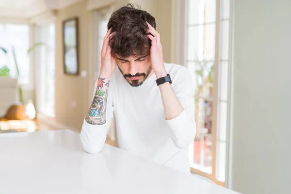 Young man wearing casual shirt sitting on white table suffering from headache desperate and stressed because pain and migraine. Hands on head.