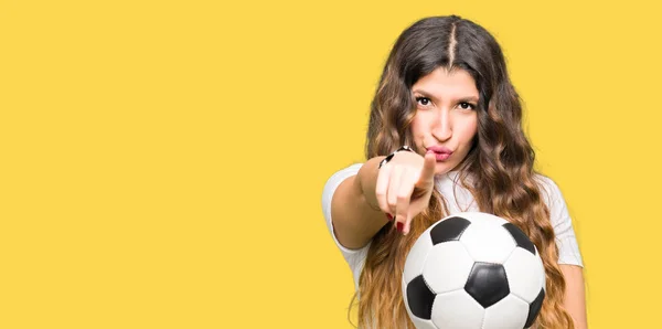 Young adult woman holding soccer football ball pointing with finger to the camera and to you, hand sign, positive and confident gesture from the front