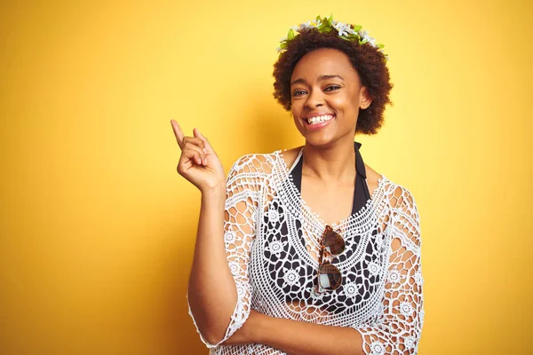 Young african american woman with afro hair wearing flowers crown over yellow isolated background with a big smile on face, pointing with hand and finger to the side looking at the camera.