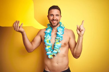Yakışıklı gömleksiz adam hawaiian lei sarı arka plan üzerinde konuşma balonu tutan giyen çok mutlu yan el ve parmak ile işaret