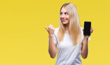 Genç güzel sarışın kadın Smartphone işaret ve başparmak kadar mutlu yüz gülümseyen tarafı ile gösteren izole arka plan üzerinde gösterilen