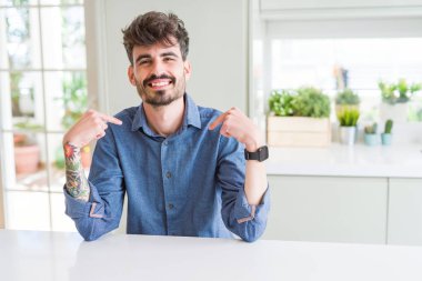 Beyaz masanın üzerinde oturan rahat gömlek giyen genç adam yüzünde gülümseme ile emin görünümlü, parmakları gururlu ve mutlu kendini işaret.