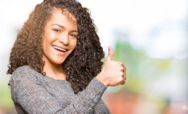 Kıvırcık saçlı, gri kazak giyen, gururlu, gülümseyen, baş parmaklarını kaldıran güzel bir kadın.