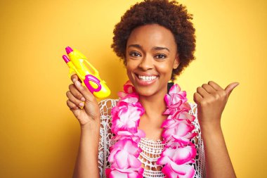 Afro saçlı genç afro-amerikalı kadın çiçek hawaiian lei ve su tabancası giyen işaret ve mutlu yüzü gülümseyerek yan başparmak ile gösteren
