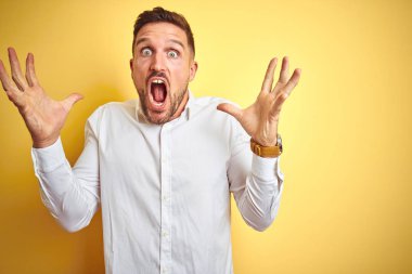 Genç yakışıklı adam sarı izole arka plan üzerinde zarif beyaz gömlek giyen deli kutluyor ve kollarını kaldırdı ve açık gözleri heyecanlı çığlık ile başarı için hayran. Kazanan kavramı