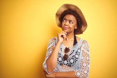 Afro saçlı genç afro-amerikalı kadın beyaz izole arka plan üzerinde yaz şapkası giyen çene üzerinde el soru, dalgın ifade düşünme. Düşünceli bir yüzle gülümsüyor. Şüphe kavramı.