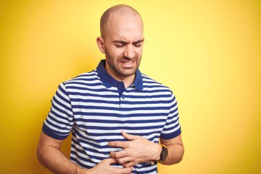 Sakallı genç kel adam mide üzerinde el ile sarı izole arka plan üzerinde rahat çizgili mavi t-shirt giyiyor çünkü hazımsızlık, ağrılı hastalık duygusu. Ağrı kavramı.