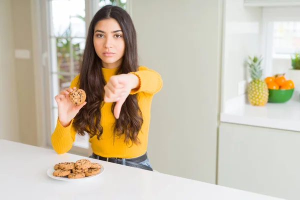 怒った顔で自宅でチョコチップクッキーを食べる若い女性 親指を下にして嫌いを示す否定的なサイン 拒絶概念 — ストック写真