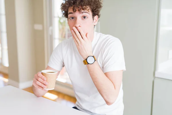Young man drinking take away cup of coffee at home cover mouth with hand shocked with shame for mistake, expression of fear, scared in silence, secret concept