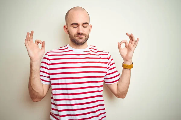 年轻的秃头男子 留着胡须 穿着白色浅条纹红色T恤 在白色孤立的背景上放松 面带微笑 闭上眼睛用手指做冥想手势 瑜伽概念 — 图库照片