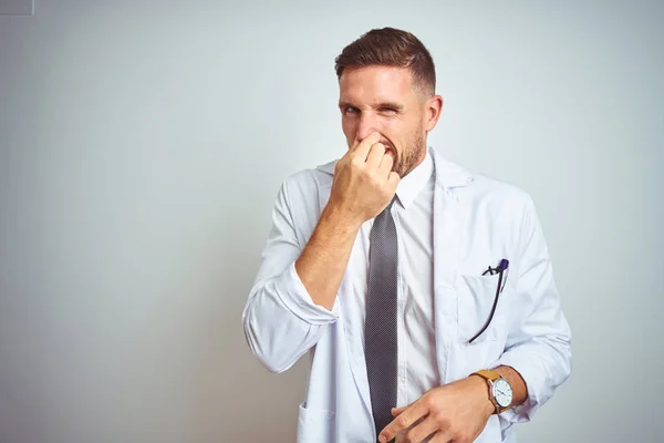 孤立した背景の上に白いプロフェショナルコートを着た若いハンサムな医者の男は 鼻に指で息を保持し 臭いと嫌な 耐えぎの良い匂いを嗅ぐ 悪臭の概念 — ストック写真