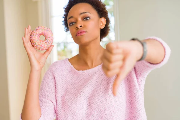 年轻的非洲裔美国妇女吃粉红色的糖甜甜圈与愤怒的脸 负面的迹象显示不喜欢与拇指向下 拒绝概念 — 图库照片