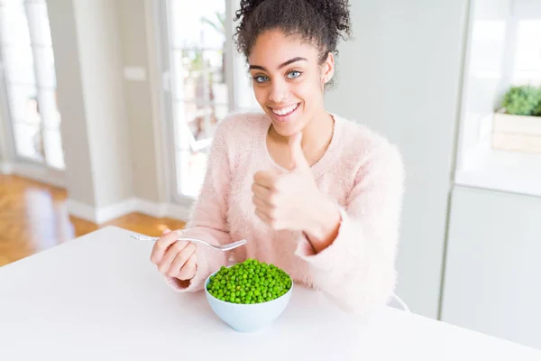 Young african american girl eating healthy green peas happy with big smile doing ok sign, thumb up with fingers, excellent sign