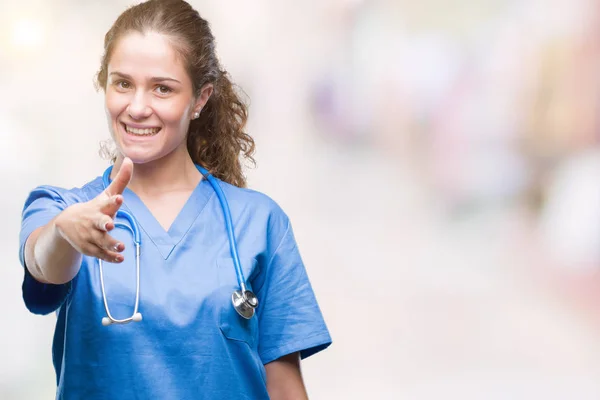 Young brunette doctor girl wearing nurse or surgeon uniform over isolated background smiling friendly offering handshake as greeting and welcoming. Successful business.