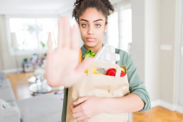 Young african american girl holding paper bag of groceries from supermarket with open hand doing stop sign with serious and confident expression, defense gesture