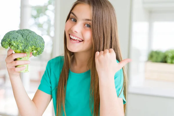 美しい若い女の子が新鮮なブロッコリーを食べて 幸せな顔の笑顔で横に親指で示しています — ストック写真