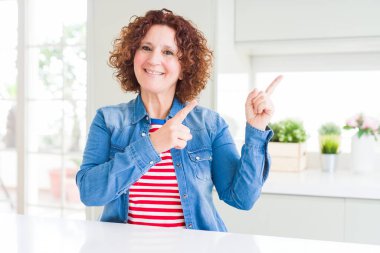 Evde kot ceket giyen kıvırcık saçlı orta yaş yaşlı kadın gülümseyerek ve yan iki el ve parmakları ile işaret kamera bakıyor.