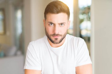 Ev şüpheci ve sinir, çapraz kolları ile yüz üzerinde onaylamaz ifade rahat beyaz t-shirt giyen genç yakışıklı adam. Negatif kişi.