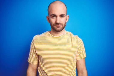 Yüzünde ciddi ifade ile mavi izole arka plan üzerinde rahat t-shirt giyen sakallı genç kel adam. Kameraya basit ve doğal bir bakış.