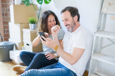 Orta yaş üst düzey romantik çift aşk etrafında kutuları ile daire katında oturan ve akıllı telefon yeni bir eve taşımak için mutlu gülümseyen kullanarak