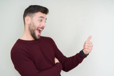 Genç yakışıklı adam izole arka plan üzerinde bir kazak giyen gururlu arıyorsunuz, yan jest kadar başparmak yapıyor gülümseyerek